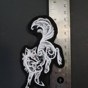 May include: White embroidered patch of a stylized wolf on a black felt background. The wolf is curled up with its tail over its back. The patch is approximately 2 inches wide.