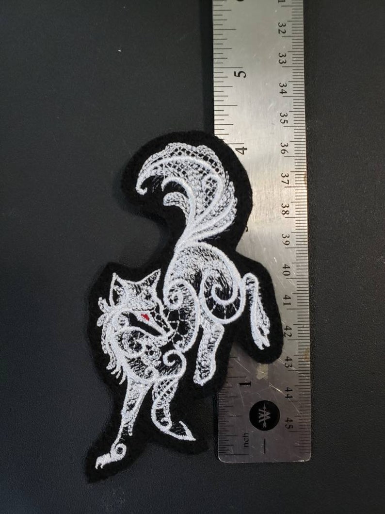 May include: White embroidered patch of a stylized wolf on a black felt background. The wolf is curled up with its tail over its back. The patch is approximately 2 inches wide.