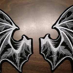 May include: A pair of black and white embroidered iron-on patches in the shape of wings. The wings have a detailed design with a white outline and black fill.