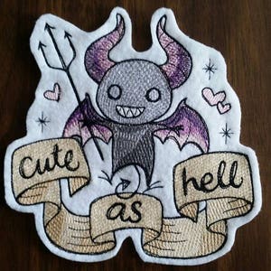 May include: Embroidered patch featuring a cute cartoon devil with purple wings and horns, holding a trident. The patch has a banner that reads "Cute as hell".