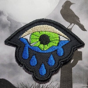 May include: An embroidered patch with a green eye with a black pupil, surrounded by a white iris. The eye is crying blue tears, with three drops falling from the eye.