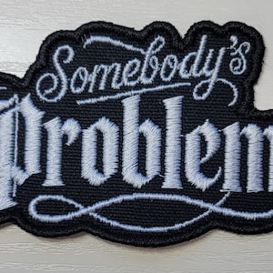 May include: A black embroidered patch with a white stitched border and the text Somebody's Problem in a stylized script and serif font. The embroidery features a black background with white thread detailing the lettering and decorative flourishes.