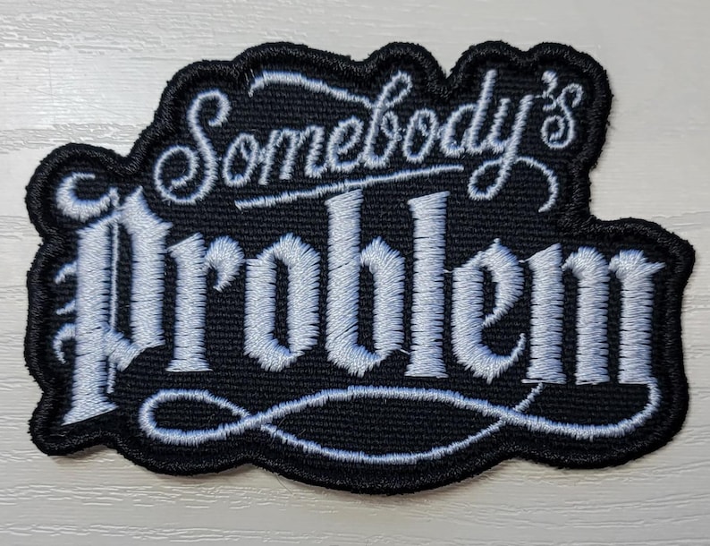 Peut inclure: Un &eacute;cusson brod&eacute; noir avec une bordure cousue blanche et le texte Somebody's Problem dans une police de caract&egrave;res stylis&eacute;e script et serif. La broderie pr&eacute;sente un fond noir avec du fil blanc d&eacute;taillant le lettrage et les fioritures.