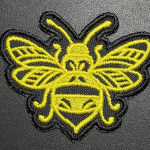 May include: Embroidered patch featuring a stylized yellow bee with black outlines. The bee has detailed wings and body segments, with a black border. The patch is designed for application on clothing or accessories.