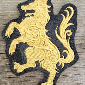 May include: An embroidered patch featuring a gold-colored wolf in a rampant position with intricate swirling patterns throughout its body, set against a solid black fabric background with a die-cut border following the shape of the wolf.