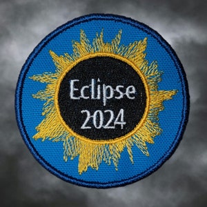 May include: A blue circular patch with a yellow sunburst design and a black circle in the center. The text "Eclipse 2024" is written in white on the black circle.