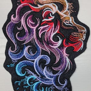 May include: Embroidered lion head patch with a colorful, swirling design. The lion's mane is a mix of pink, purple, blue, and teal. The patch is on a black felt background.