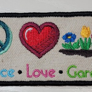 May include: A rectangular embroidered patch on a beige fabric base with a black border. It features a teal peace sign, a red heart, and a small garden scene with blue, yellow, and red flowers. Below, the text reads Peace, Love, Garden.