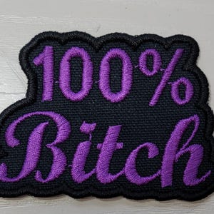 May include: Black embroidered patch with the words "100% Bitch" in purple script. The patch has a black scalloped border and is likely designed to be sewn or ironed onto clothing or accessories.