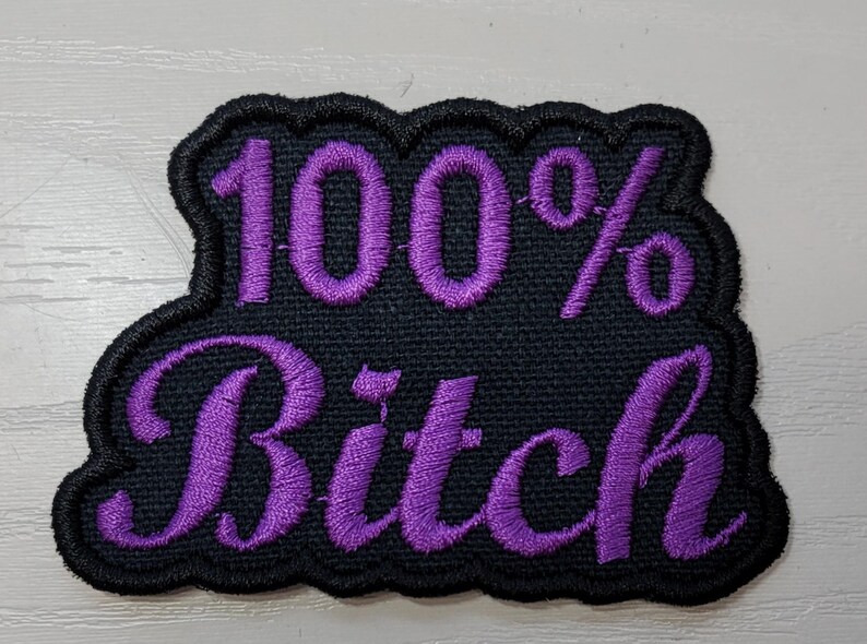 May include: Black embroidered patch with the words "100% Bitch" in purple script. The patch has a black scalloped border and is likely designed to be sewn or ironed onto clothing or accessories.