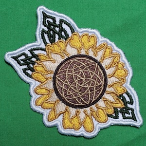 May include: A white and brown embroidered patch of a sunflower with a Celtic knot design in the center. The sunflower has yellow petals and a brown center. The patch is surrounded by green leaves.