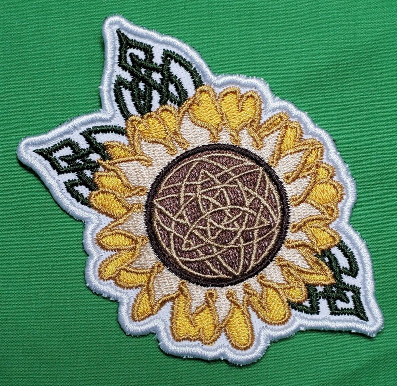 Pode incluir: Um remendo bordado branco e marrom de um girassol com um design de n&oacute; celta no centro. O girassol tem p&eacute;talas amarelas e um centro marrom. O remendo &eacute; cercado por folhas verdes.