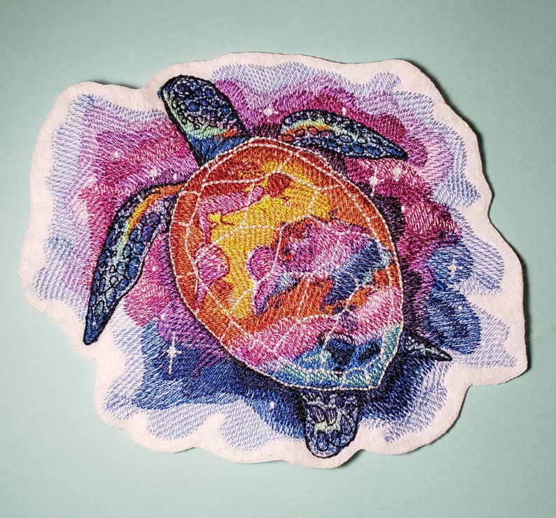 May include: A colorful embroidered patch of a sea turtle with a galaxy design. The turtle is a vibrant mix of pink, orange, yellow, and blue.