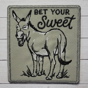 May include: A gray embroidered patch with a cartoon donkey and the text "Bet Your Sweet" in black cursive lettering.