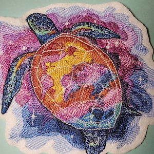 May include: Embroidered patch of a colorful sea turtle swimming in a galaxy of stars. The turtle is a vibrant orange, yellow, blue, and purple. The background is a swirling mix of blue, purple, and pink.