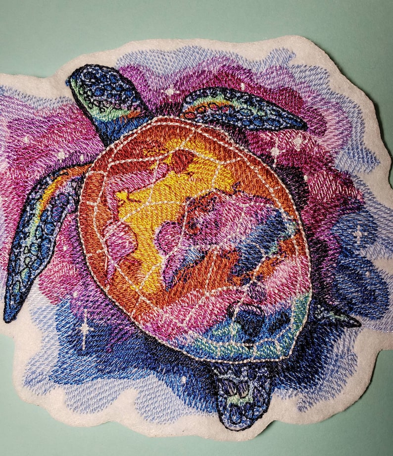 May include: Embroidered patch of a colorful sea turtle swimming in a galaxy of stars. The turtle is a vibrant orange, yellow, blue, and purple. The background is a swirling mix of blue, purple, and pink.