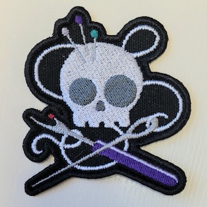 May include: Embroidered patch featuring a skull with gray eyes and colorful pins in its head. Crossed needles and a purple measuring tape are below the skull. The patch has a black border and is on a white surface.
