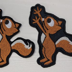 Peut inclure: Deux patchs brod&eacute;s thermocollants repr&eacute;sentant un &eacute;cureuil de dessin anim&eacute; les bras lev&eacute;s en l'air. L'&eacute;cureuil est brun avec un ventre et une queue blancs. Les patchs sont sur un fond noir.