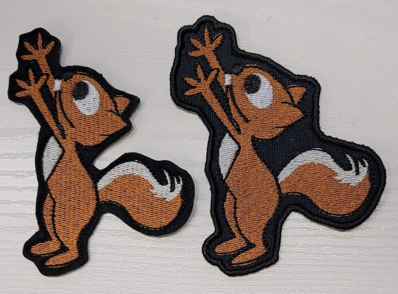 Peut inclure: Deux patchs brod&eacute;s thermocollants repr&eacute;sentant un &eacute;cureuil de dessin anim&eacute; les bras lev&eacute;s en l'air. L'&eacute;cureuil est brun avec un ventre et une queue blancs. Les patchs sont sur un fond noir.