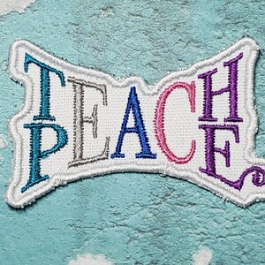 May include: A white fabric patch with the word "TEACH" embroidered in colorful thread. The letters are outlined in white and the word is in a stylized font. The letters are blue, gray, blue, pink, and purple.