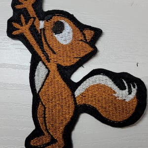Peut inclure: Patch brod&eacute; thermocollant repr&eacute;sentant un &eacute;cureuil de dessin anim&eacute; brun avec des yeux blancs et une queue blanche, debout sur ses pattes arri&egrave;re, les bras lev&eacute;s en l'air.