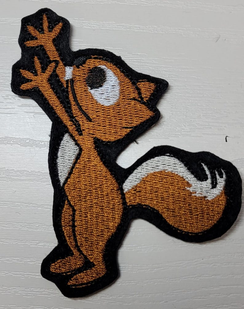 Peut inclure: Patch brod&eacute; thermocollant repr&eacute;sentant un &eacute;cureuil de dessin anim&eacute; brun avec des yeux blancs et une queue blanche, debout sur ses pattes arri&egrave;re, les bras lev&eacute;s en l'air.