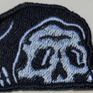 May include: Embroidered patch featuring a stylized skull design in light blue and black. The skull is framed by a dark blue border, with detailed stitching creating a textured appearance. The patch is likely for clothing or accessories.