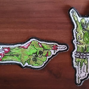 May include: Two embroidered iron-on patches featuring zombie hands. One patch shows a green zombie hand giving the 'rock on' sign. The other patch shows a green zombie hand pointing with bloody dripping fingers.