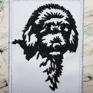 May include: Black and white embroidered patch of a dog's head with a long, shaggy beard.