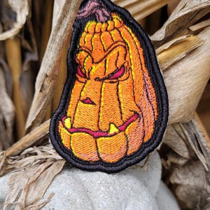 K&ouml;nnte beinhalten: Ein gestickter Aufn&auml;her mit einem stilisierten orangefarbenen K&uuml;rbis mit einem dunklen, geschnitzten Gesichtsausdruck, einschlie&szlig;lich schmaler roter Augen und gelber Z&auml;hne. Das Design ist mit dickem schwarzem Garn umrandet, das die Rippen und den Stiel betont.