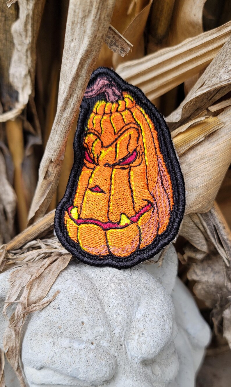 K&ouml;nnte beinhalten: Ein gestickter Aufn&auml;her mit einem stilisierten orangefarbenen K&uuml;rbis mit einem dunklen, geschnitzten Gesichtsausdruck, einschlie&szlig;lich schmaler roter Augen und gelber Z&auml;hne. Das Design ist mit dickem schwarzem Garn umrandet, das die Rippen und den Stiel betont.