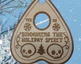May include: Wooden Christmas ornament shaped like a Ouija board with the words "Summoning the Holiday Spirit" and two skulls on either side of a Christmas tree. The ornament has a hole at the top for hanging.