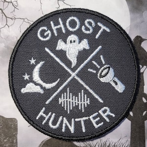 May include: A black and white embroidered patch with the text "Ghost Hunter" in a circle. The design includes a ghost, a crescent moon, a cloud, a flashlight, and a series of vertical lines.