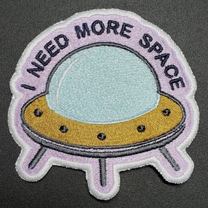 May include: A purple and white embroidered patch with a cartoon spaceship and the text "I NEED MORE SPACE".