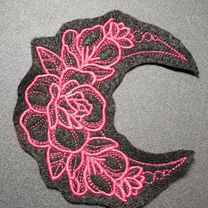 May include: A dark gray, crescent-shaped embroidered patch featuring a vibrant pink floral design. The patch showcases a detailed rose and leaf pattern, with delicate stitching outlining the flowers and leaves. The patch is likely for decorative purposes.