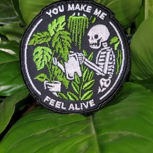 May include: A black and white embroidered patch with a skeleton watering plants. The patch has the text "You make me feel alive" around the edge.