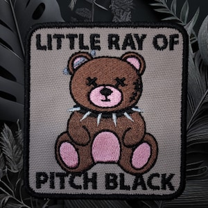 May include: A brown teddy bear with a spiked collar and pink paws. The bear is sitting on a beige background with the text "Little Ray of Pitch Black" in black.