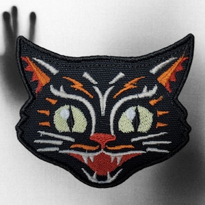 May include: Black embroidered cat patch with orange and white details. The cat has a menacing expression with sharp teeth and large eyes.