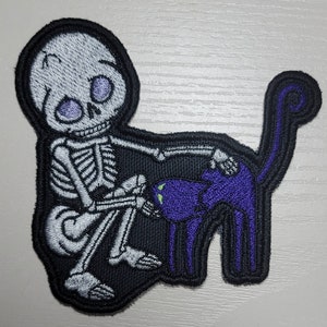 May include: Embroidered patch featuring a white skeleton petting a purple cat. The skeleton has purple eyes, and the cat has green eyes. The patch is outlined in black.