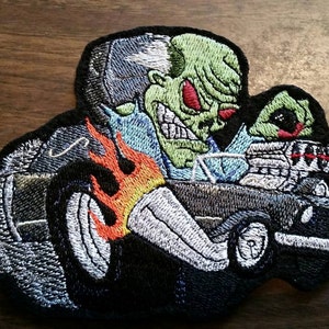 Peut inclure: Patch brod&eacute; repr&eacute;sentant un monstre vert conduisant une voiture de course noire avec des flammes sortant de l'&eacute;chappement. Le monstre a des yeux rouges et un sourire mena&ccedil;ant.