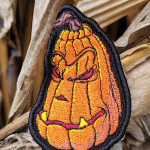 K&ouml;nnte beinhalten: Ein gestickter Aufn&auml;her in Form eines stilisierten orangefarbenen K&uuml;rbisses mit einem grimmigen Gesichtsausdruck, spitzen gelben Z&auml;hnen und dunkelroten Augen. Das Design ist mit dickem schwarzem Garn umrandet, das die Konturen betont.