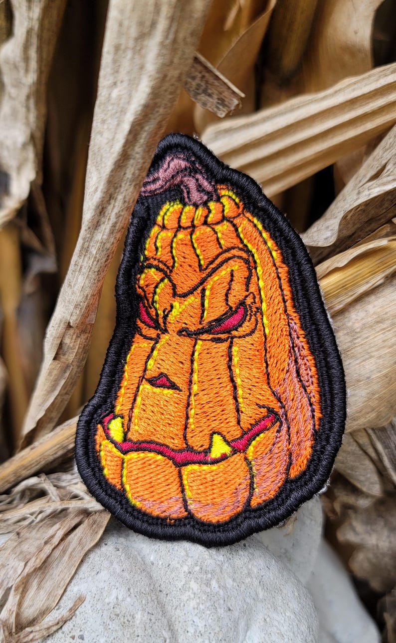 K&ouml;nnte beinhalten: Ein gestickter Aufn&auml;her in Form eines stilisierten orangefarbenen K&uuml;rbisses mit einem grimmigen Gesichtsausdruck, spitzen gelben Z&auml;hnen und dunkelroten Augen. Das Design ist mit dickem schwarzem Garn umrandet, das die Konturen betont.
