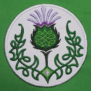 May include: A white embroidered patch with a green Celtic knot thistle design. The thistle is surrounded by green leaves and has a purple flower.