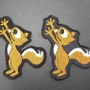Peut inclure: Deux patchs brod&eacute;s &agrave; repasser repr&eacute;sentant un &eacute;cureuil de dessin anim&eacute; les bras lev&eacute;s en l'air. L'&eacute;cureuil est brun avec une queue blanche et un contour noir. Les patchs sont sur un fond gris.