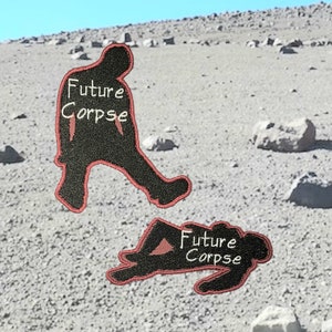 May include: Two black embroidered patches with red stitching. The patches are shaped like a person. One patch shows a person walking with the text "Future Corpse" written on the chest. The other patch shows a person lying down with the text "Future Corpse" written on the chest.