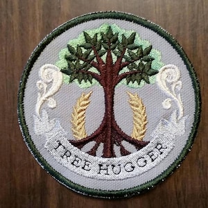 Puede incluir: Un parche redondo bordado de color gris con un árbol verde, raíces marrones y espigas de trigo doradas. El texto "TREE HUGGER" está escrito en blanco sobre una pancarta blanca.