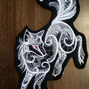 May include: White embroidered wolf patch with intricate details on a black fabric background. The wolf's eye is a bright red.