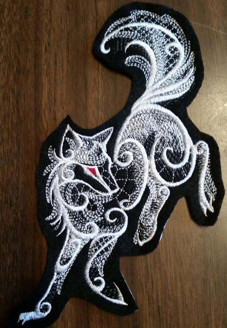 May include: White embroidered wolf patch with intricate details on a black fabric background. The wolf's eye is a bright red.