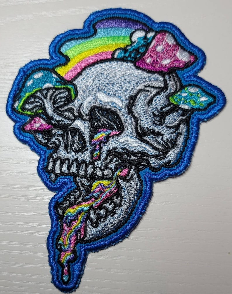 Op de afbeelding: Een blauwe geborduurde patch met een witte schedel, een regenboog en kleurrijke paddenstoelen. De schedel heeft een regenboog die uit zijn mond druipt.