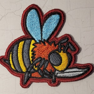 May include: A red, yellow, black, and blue embroidered patch of a cartoon bee holding a silver knife. The bee has a red and orange fuzzy head and black eyes.